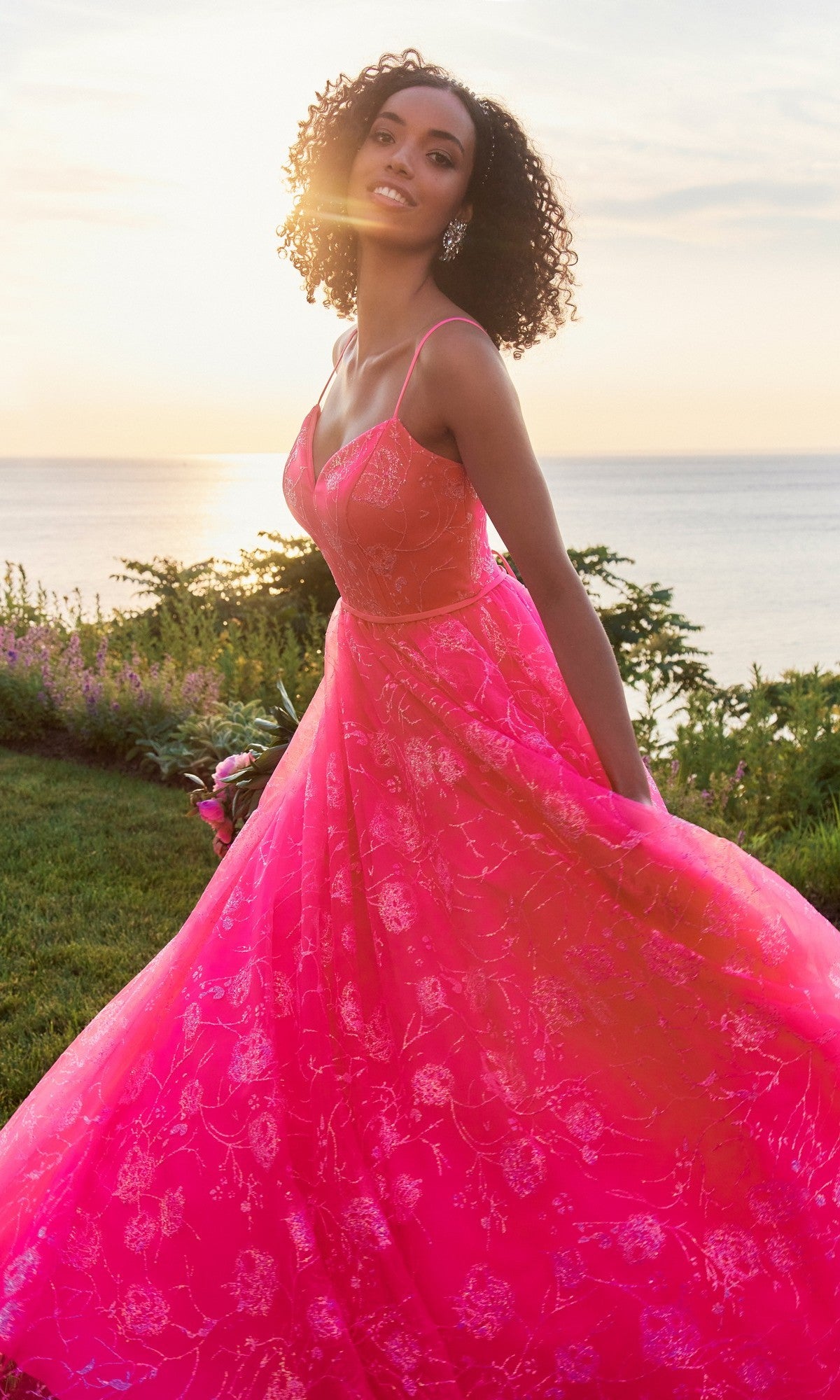 A woman in a Morilee 49153 Long Prom Dress in Pink Panther stands outdoors by the water at sunset, holding her gown and smiling.